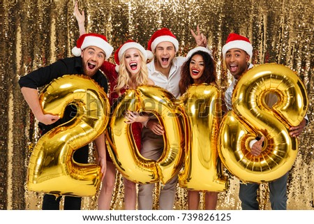 Group of smiling friends celebrating New Year while standing with golden balloon 2018 numbers and looking at camera isolated over shiny golden background