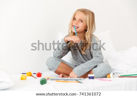 Happy blonde girl with pencil in her mouth sitting in bed, looking at camera