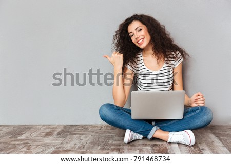 Nice woman with beautiful smile sitting in lotus pose on the floor with silver computer on legs gesturing thumb aside, submitting something copy space