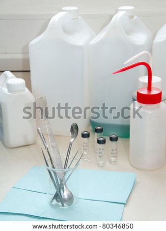 Research lab: desk with scientific instruments, spoons, pair of pincers, test-tubes and large plastic flasks in the background