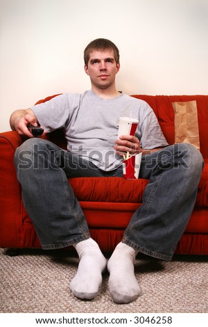 negative side of american lifestyle - a bored young man on a couch with a tv remote and junk food