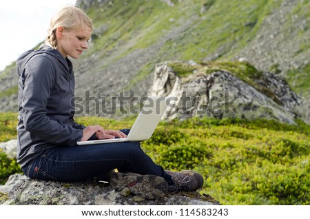 young blond woman sits with a laptop on a stone before mountain landscape