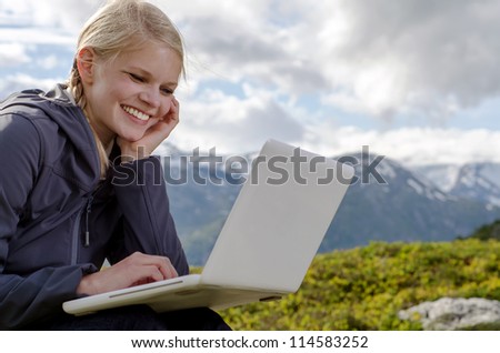 young blond woman sits with a laptop before a mountain landscape