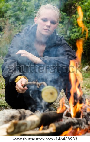 a blond young woman sits at the campfire and grills vegetables