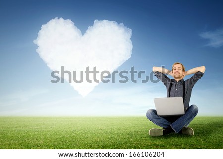a man sits relaxed with laptop on the meadow with a cloud in heart shape in the background