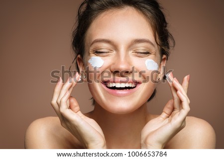 Laughing girl applying moisturizing cream on her face. Photo of young girl with flawless skin on beige background. Skin care and beauty concept