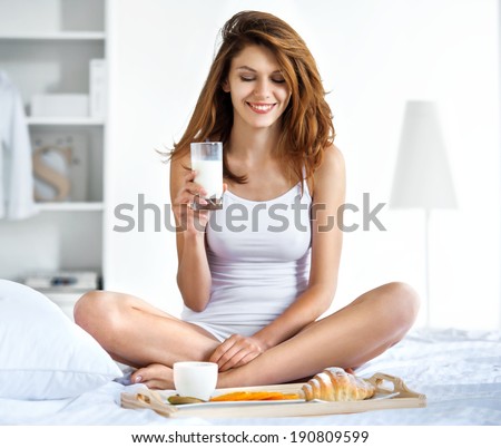 It is okay to drink a glass of milk before going to sleep? / smiling brunette woman hold a glass with milk while sitting on the bed
