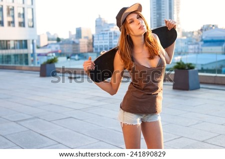 Sexy swag girl is standing on the street holding skateboard behind the back. The girl in joyful feelings / outdoors photography of young Caucasian woman on the street