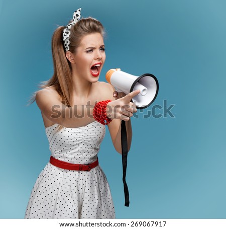 Angry pin-up girl shouting into a megaphone, mouthpiece, speaking trumpet. Film making or film production concept / photo set of young American pin-up model on blue background