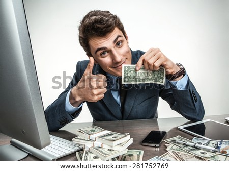 Cheerful smiling man showing thumbs up success sign / modern businessman at his desk with computer and a lot of money