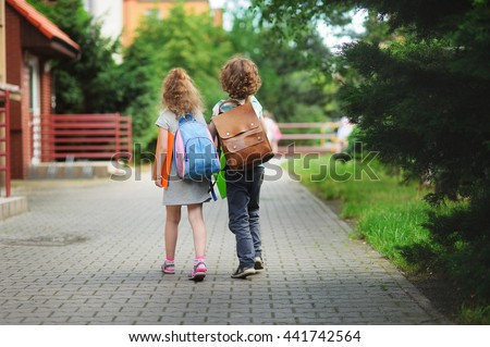 Two young students go to school hand in hand. Children behind shoulders have satchels. These are pupils of elementary school. Back to school. Lovely first graders.