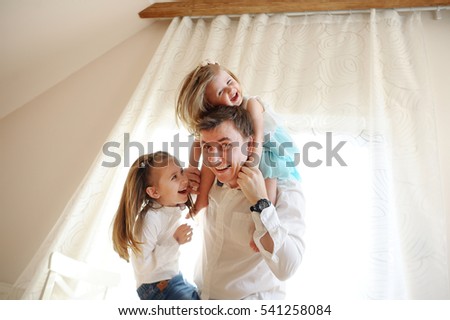 The young man cheerfully spends time with the little daughters. Younger girl sits at the father on shoulders, another trying to climb the dad. Children and father delighted with a play.