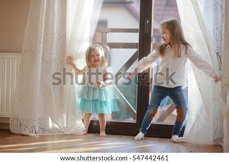 Two little sisters play in the room. Girls hide behind curtains, and then joyfully jump out because of curtains.