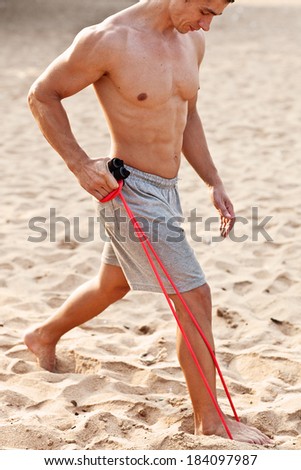 Young athletic man exercising and doing fitness with a chest expander(resistance band ) on the beach