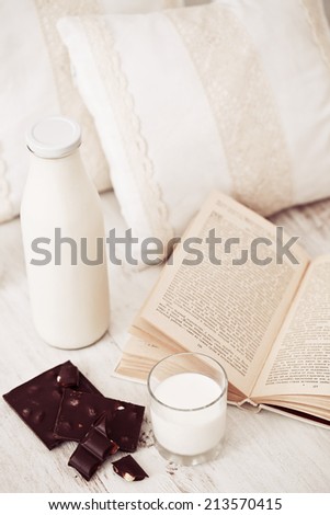 Still life interior details, glass of milk, chocolate and a book near white cozy pillow