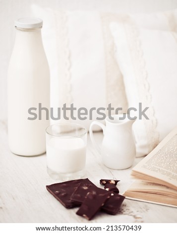 Still life interior details, glass of milk, chocolate and a book near white cozy pillow