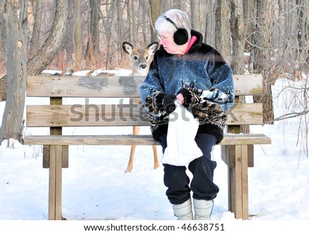 An elderly woman sitting on a park bench with a deer looking at her from behind.