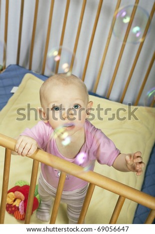 little baby holds on to the baby bed and looks behind the bubbles