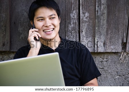A happy asian student talking on the phone while working on his laptop