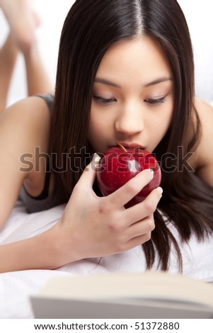 A shot of a beautiful asian college student studying on her bed and eating an apple