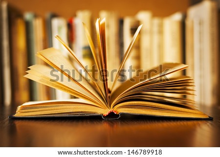 Hardcover book lying open on its cover with the pages fanned above it and a row of books on a bookshelf visible behind, shallow dof