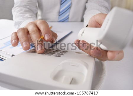 Close up view of the hands of a man making a telephone call on a landline holding the handset close to the camera and dialling in the number on the keypad with the other hand