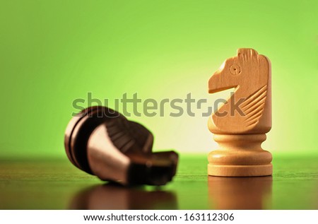 Two wooden knights in a game of chess with the darker piece lying on its side and the lighter one upright, low angle view on green
