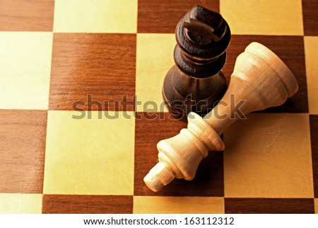 Close up overhead view of two wooden chess pieces on a chessboard with the lighter piece lying on its side and the black king standing upright