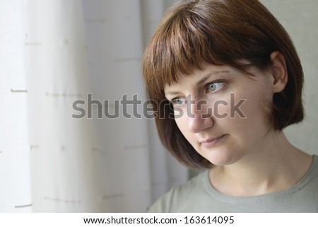 Close up head portrait of an attractive serious young woman standing staring out of a window with a pensive expression