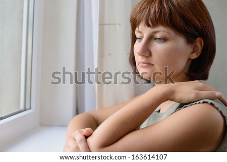Sad young woman standing thinking with with downcast eyes, a serious expression and her arm resting on a windowsill