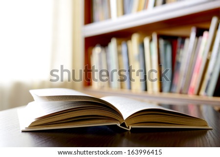 Close-up of an open book , on a wooden table, with bookshelvClose-up of an open book , on a wooden table, with bookshelves in the background, as an invitation to study literaturees in the background,