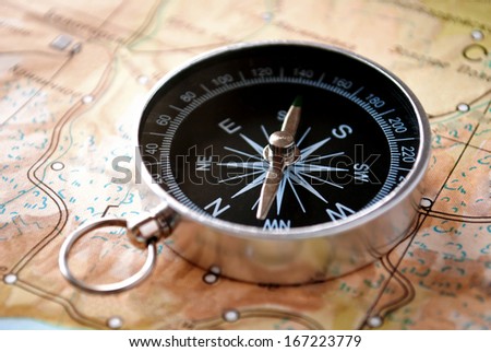 Handheld compass lying on a map showing the needle and cardinal points of north, south, east and west to aid in magnetic navigation to plot a route or direction to a specific destination