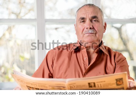 Friendly elderly man with a moustache sitting reading a newspaper in front of a bright airy window looking at the camera with a smile