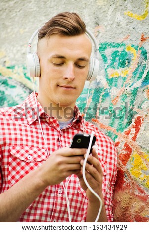 Happy handsome man enjoying his music sitting on the ground against a graffiti covered wall looking up tunes on his MP3 player