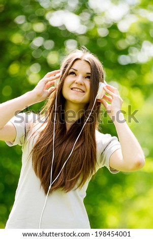 Young woman concentrating on her music standing in a leafy green wooded park holding her headphones to her ears with her head tilted back looking up with a contented expression