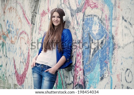 Trendy young woman with long brunette hair leaning against a grungy graffiti covered wall listening to music on her headphones