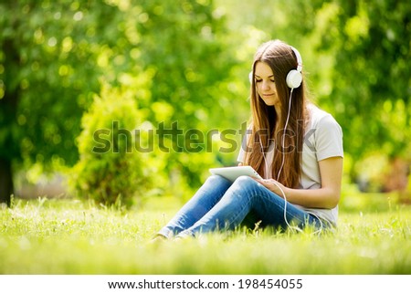 Young woman listening to music in a lush park sitting on the grass with her tablet on her knees selecting a tune to listen to on her headphones