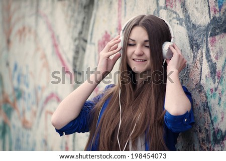 Attractive teenage girl enjoying her music standing against a graffiti covered wall listening to her headphones with closed eyes and a blissful smile