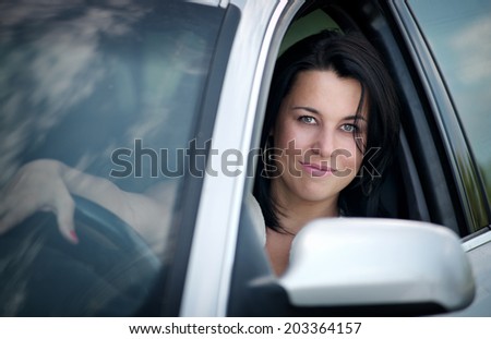 Beautiful young woman driving a car looking through the open side window at the camera with a lovely friendly smile