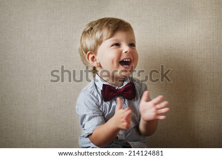 Beautiful little boy wearing a stylish maroon bow tie laughing and clapping his hands against a grey wall with vignetting