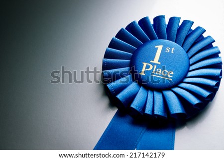 Blue first place winner rosette or badge from pleated ribbon with central text to be awarded to the winner of a competition on a graduated grey background with copyspace