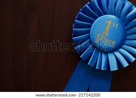 First place winners rosette or badge in pleated blue ribbon with central text to be awarded as a prize in a competition, race, or sport at an oblique angle on grey with copyspace