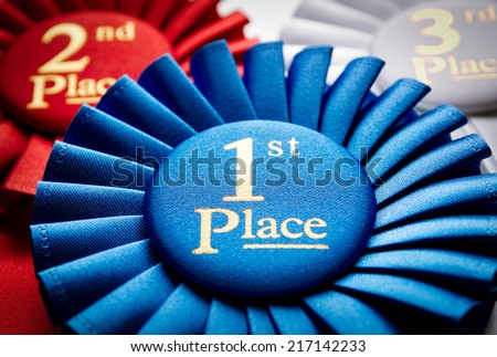 1st place blue winners rosette or badge to be awarded as a prize to the winner of a competition made of pleated blue ribbon with central text in gold with a 2nd place red rosette behind
