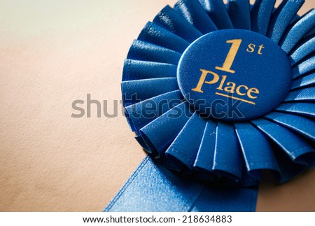 Blue first place winner rosette or badge from pleated ribbon with central text to be awarded to the winner of a competition on a graduated beige background with copyspace