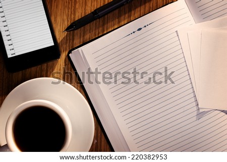 Open notebook with blank white lined pages with a cup of black coffee alongside a phone on a wooden desk, overhead view