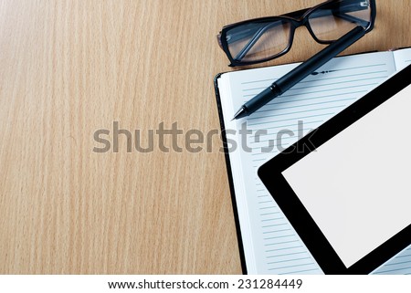 Modern tablet PC displaying a white page next to eyeglasses and a pen on a classical open agenda or notebook with empty pages, on a wooden table or desk