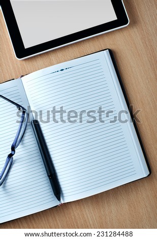 Modern tablet PC displaying a white page next to eyeglasses and a pen on a classical open agenda or notebook with empty pages, on a wooden table or desk