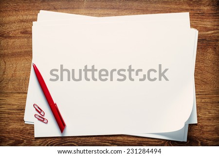 Red ballpoint pen and paperclips lying on a stack of blank sheets or pages of white paper with copyspace for your text on a wooden desk, view from above
