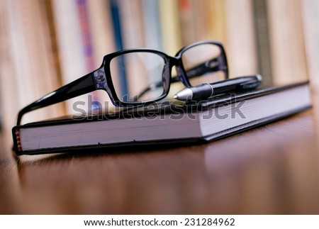 Close up Eyeglasses with Black Frame and Black Ballpoint Pen on Top of the Book at the Office Table.