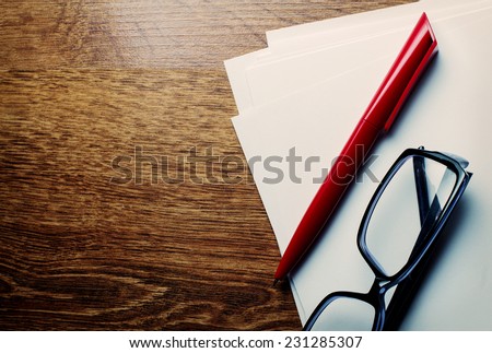 Red ballpoint pen and and a pair of reading glasses lying on pages of blank white paper on a wooden desk with copyspace, overhead view